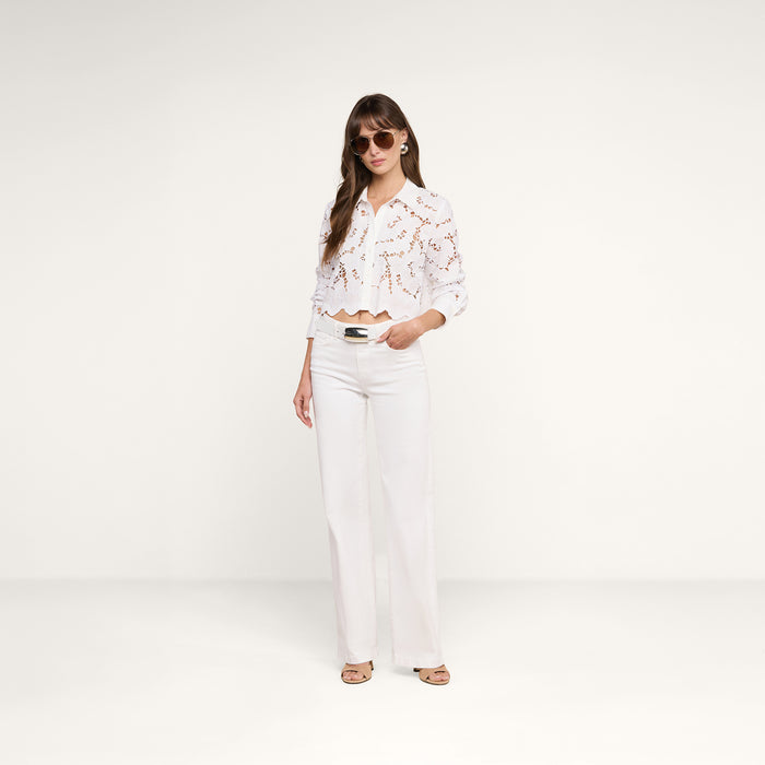 A woman wearing sunglasses and the Seychelle Cropped Eyelet Blouse— a white long-sleeve lace top with red pattern detailing— paired with white wide-leg pants and beige sandals, stands against a plain white background, effortlessly capturing relaxed beach style.