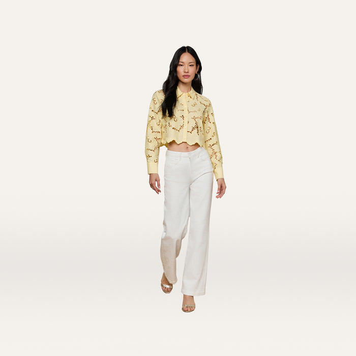 A woman walks forward wearing the Seychelle Cropped Eyelet Blouse, featuring a yellow floral eyelet design, paired with white wide-leg pants and open-toe shoes, against a plain light background.