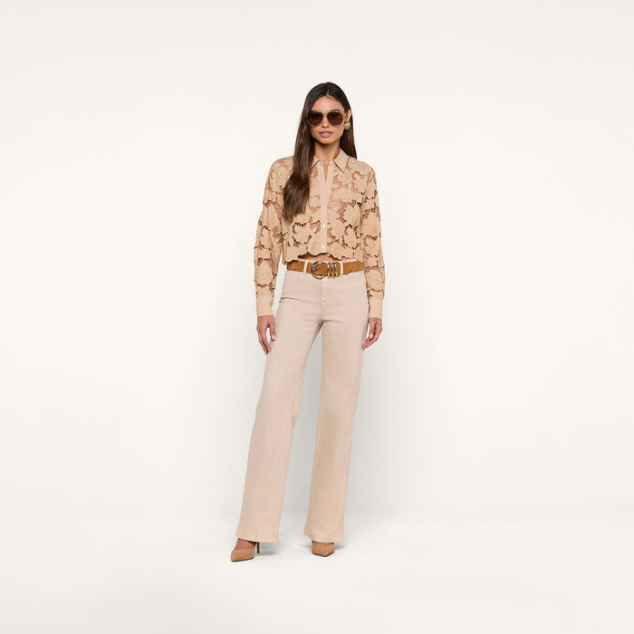 A woman stands against a white background wearing sunglasses, the Seychelle Cropped Eyelet Blouse, high-waisted light beige pants, and tan heels, with her hands in her pockets.
