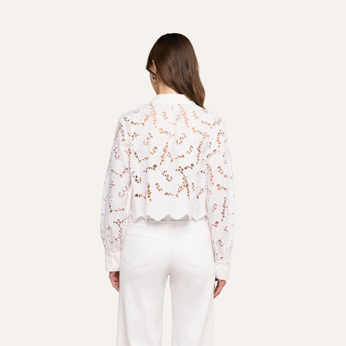 A woman stands with her back to the camera, wearing the Seychelle Cropped Eyelet Blouse—an all-white, long-sleeve top featuring a semi-sheer floral eyelet pattern and scalloped hem—paired with high-waisted white pants. Her brown hair is worn down, and the background is a plain light beige.