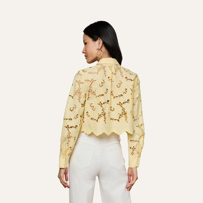 A woman with long dark hair, wearing large hoop earrings and high-waisted white pants, is shown from the back against a plain light background, modeling the Seychelle Cropped Eyelet Blouse in yellow sheer floral fabric.