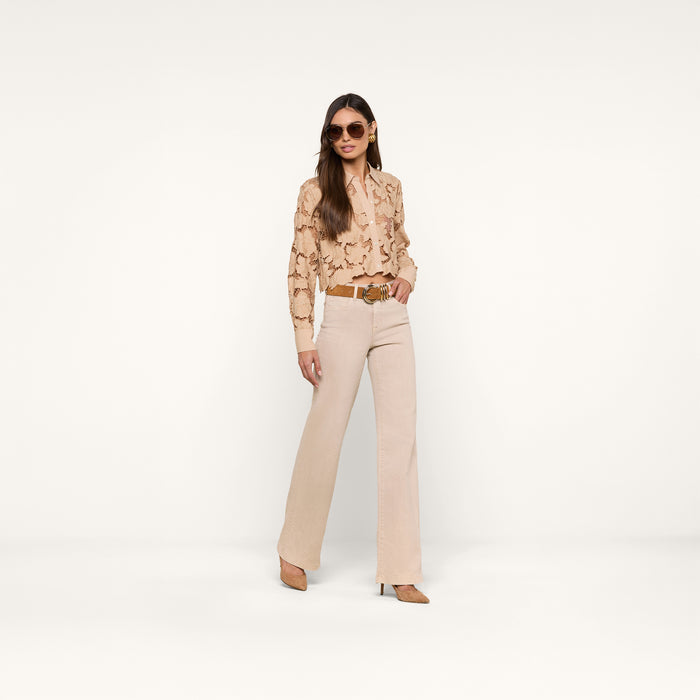 A woman stands against a plain white background, facing forward with one hand in her pocket, wearing beige trousers, a brown belt, heels, sunglasses, and the Seychelle Cropped Eyelet Blouse.