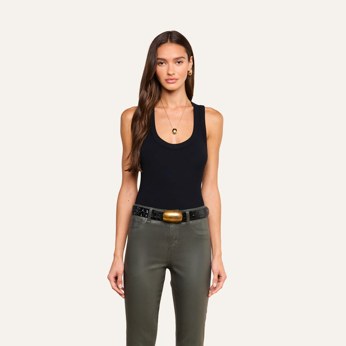 A woman with long brown hair stands facing forward against a plain light background, wearing the Shania Racerback Tank in black ribbed knit, along with olive green pants, a wide belt with a gold buckle, gold hoop earrings, and a necklace.