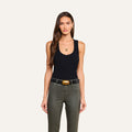 A woman with long brown hair stands facing forward against a plain light background, wearing the Shania Racerback Tank in black ribbed knit, along with olive green pants, a wide belt with a gold buckle, gold hoop earrings, and a necklace.