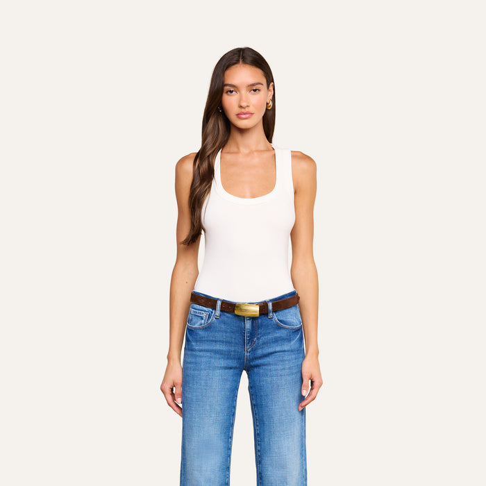 A woman with long brown hair wears the Shania Racerback Tank, styled with blue jeans featuring front pockets and a brown belt, standing against a plain light background.