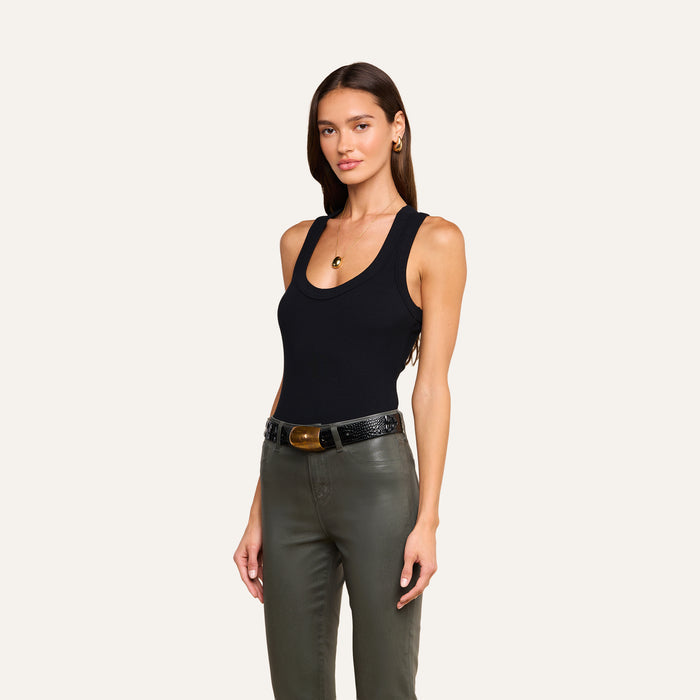 A woman with long brown hair wears the Shania Racerback Tank in black, paired with olive green pants and a black belt. She accessorizes with gold earrings and a necklace, standing against a plain light background and facing forward with a relaxed expression.
