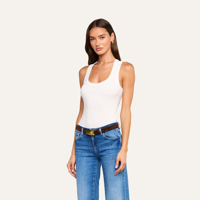 A woman with long brown hair stands against a plain light background, facing forward with one hand by her side, wearing the Shania Racerback Tank in white, paired with blue jeans and a brown belt.