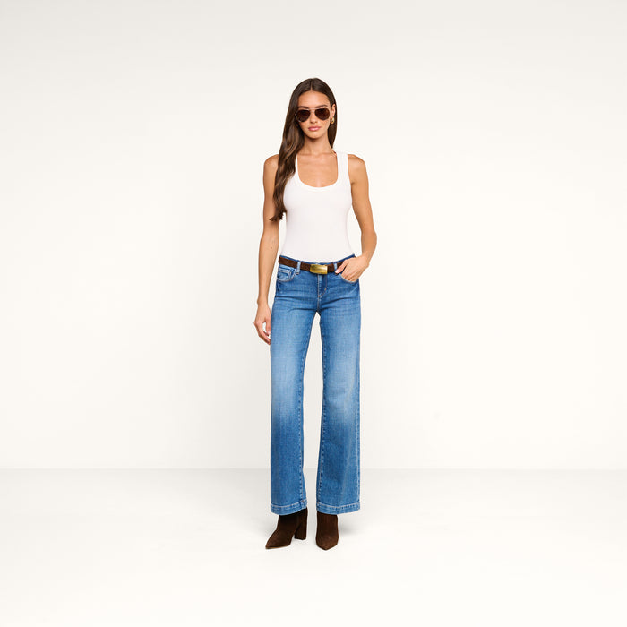 A woman stands facing forward against a plain white background, wearing sunglasses, blue jeans with brown boots, and the Shania Racerback Tank in white.