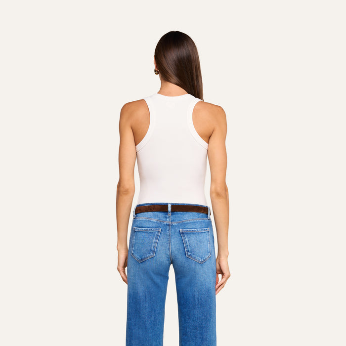 A woman stands facing away, wearing the Shania Racerback Tank in white with blue jeans and a brown belt. Her straight brown hair falls down her back against a plain light background.