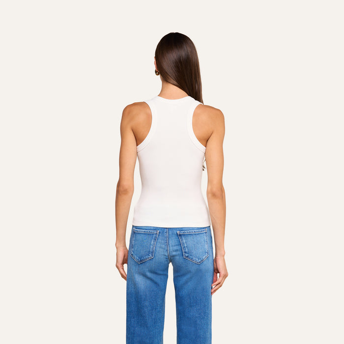 A woman stands facing away, wearing the Shania Racerback Tank in white with blue jeans against an off-white background.