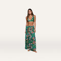 A woman stands barefoot against a plain white background, wearing the Sharon Cover-Up Pant—a chic green floral long skirt with a drawstring waist—paired with a matching bikini top. This stylish beachwear look is perfect for warm, sunny days.