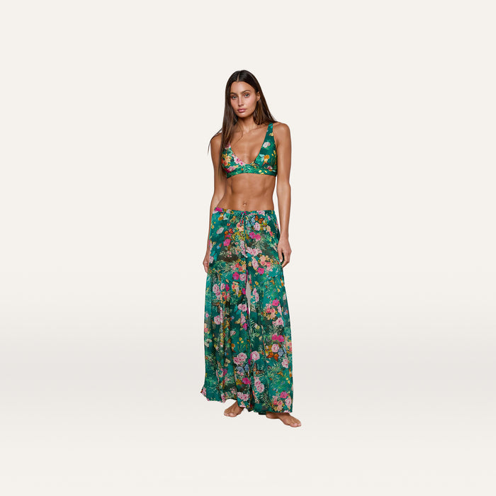 A woman stands barefoot against a plain white background, wearing the Sharon Cover-Up Pant—a chic green floral long skirt with a drawstring waist—paired with a matching bikini top. This stylish beachwear look is perfect for warm, sunny days.