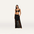 A woman stands barefoot against a plain light background, wearing a black bikini top and the Sharon Cover-Up Pant—a sheer, lacy black wide-leg style—with her left hand in her pocket.