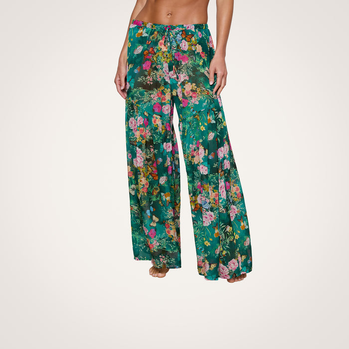 A person stands barefoot against a plain background, showcasing the lower torso and legs while wearing the Sharon Cover-Up Pant—loose, wide-leg green floral pants featuring a drawstring waist.
