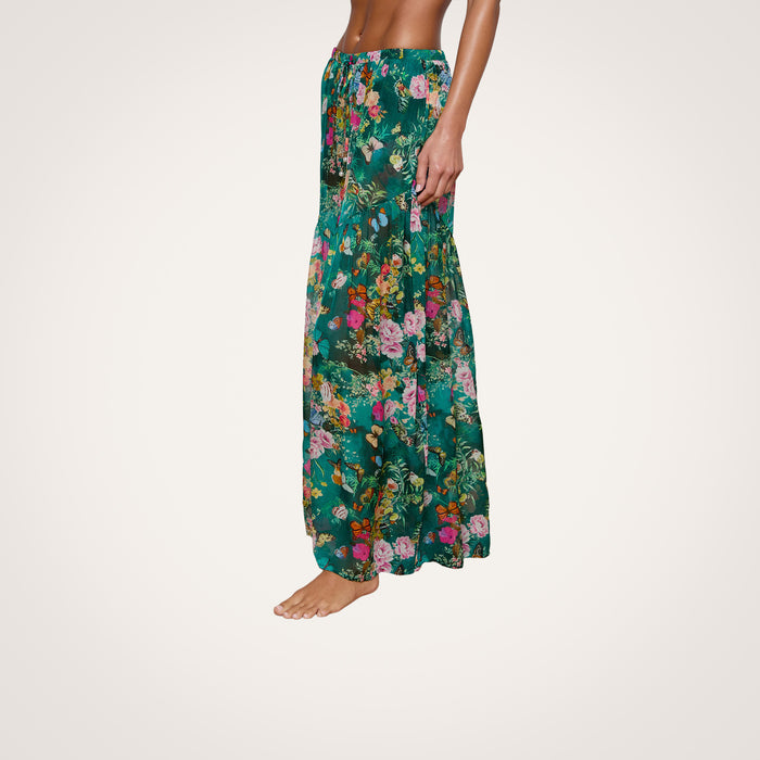 A woman stands barefoot, wearing the Sharon Cover-Up Pant—a long, green floral design with a drawstring waist. Her upper body is cropped out, and the background is plain white.
