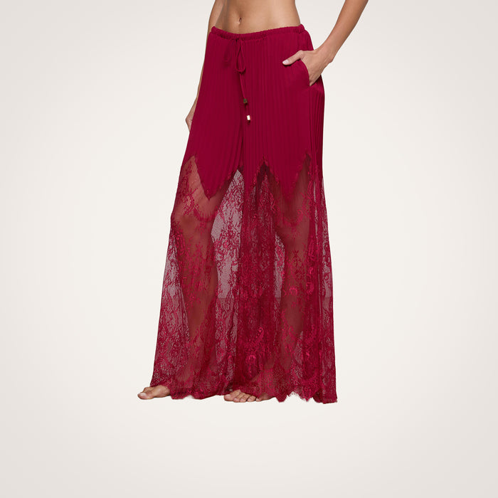 A person stands barefoot against a plain light background wearing the Sharon Cover-Up Pant, featuring sheer burgundy lace with a drawstring waist, pockets, and delicate sheer lace panels.