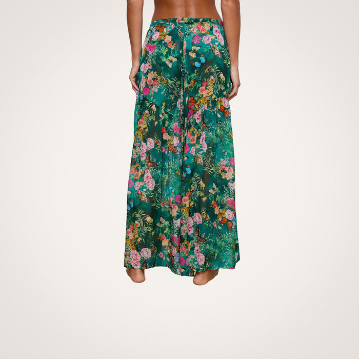 A person standing barefoot, shown from the back, is wearing the Sharon Cover-Up Pant: a pair of wide-leg, high-waisted green pants featuring a colorful floral pattern and a drawstring waist—perfect as beachwear cover-up.