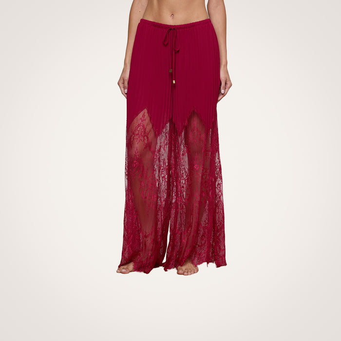 A person wearing the Sharon Cover-Up Pant—a long, sheer red lace pant with sheer lace panels and a drawstring waist, featuring solid fabric lining at the top—standing barefoot against a plain background.