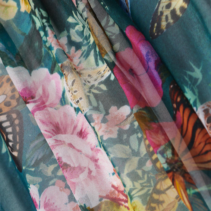 Close-up of the Sharon Cover-Up Pant, showcasing colorful floral and butterfly patterns on semi-sheer, flowing fabric beachwear with pink flowers and multicolored butterflies against a green background.