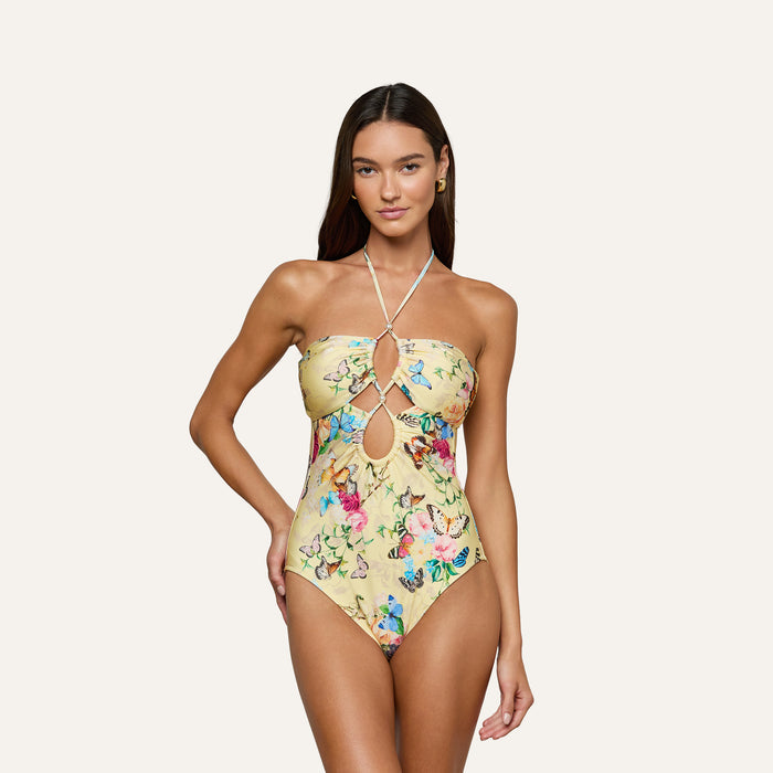A woman wearing the Shiva One-Piece Swimsuit, a strapless, halter-neck yellow swimsuit adorned with a colorful butterfly print and keyhole cutouts, stands against a plain light background.