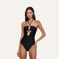 A woman modeling the Shiva One-Piece Swimsuit, featuring black fabric with keyhole cutouts and adjustable straps, stands against a plain light background.
