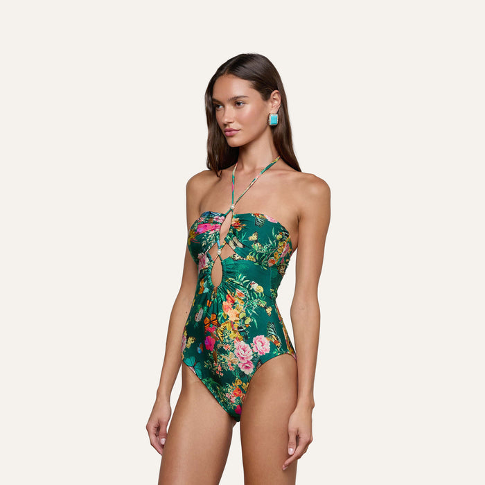 A woman wearing the Shiva One-Piece Swimsuit, featuring a green butterfly floral print, keyhole cutout details, and halter straps, stands against a plain background. She has long brown hair and is accessorized with turquoise earrings.