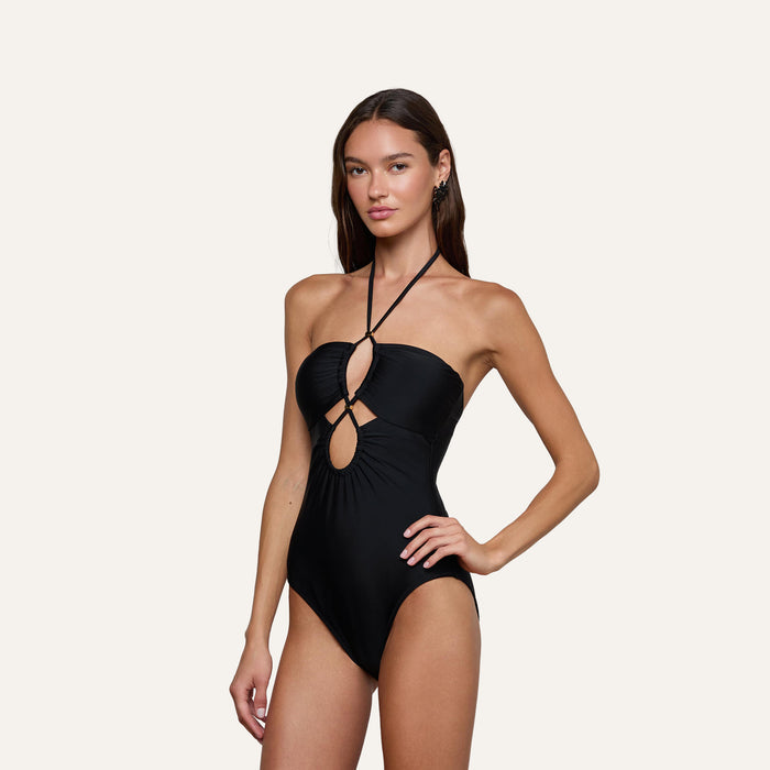 A woman poses in the Shiva One-Piece Swimsuit, featuring a black halter neck design with keyhole cutout details, against a plain light background.