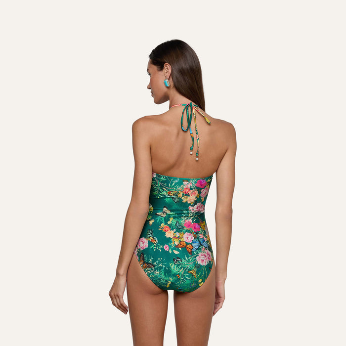 A woman stands with her back to the camera, wearing the Shiva One-Piece Swimsuit—a green butterfly floral print halter style—paired with her hair down and colorful earrings, set against a plain light background.