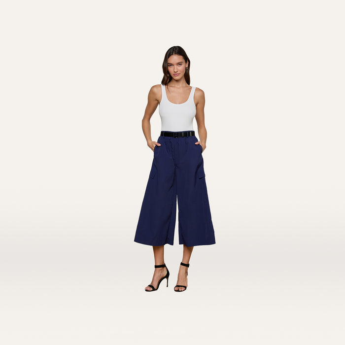 A woman stands against a plain light background wearing a white tank top, the Shoda Cropped Wide-Leg Pant in navy blue with a black belt, and black high-heeled sandals.