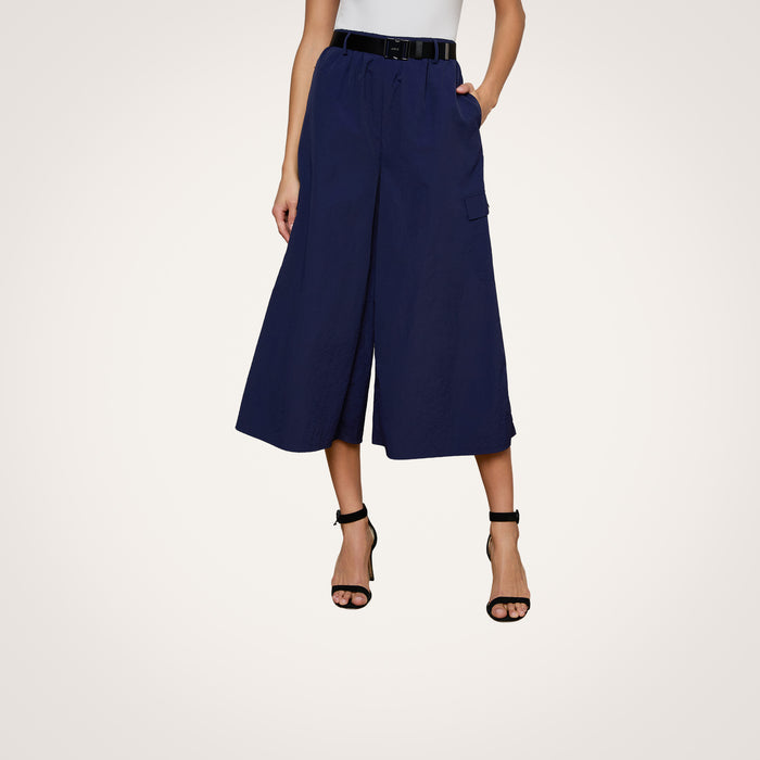A woman models the Shoda Cropped Wide-Leg Pant in navy blue, styled with a black belt and black high-heeled sandals. Her hands are tucked into the pants’ pockets, while her upper body remains out of frame against a plain white background.