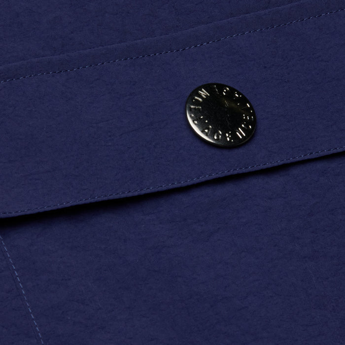 Close-up of the deep blue textured fabric from the Shoda Cropped Wide-Leg Pant, showcasing a shiny round metal snap button closure engraved with text—an element reminiscent of cargo pant detailing that secures an overlapping flap.