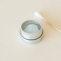 An open metal tin containing silicone grease, with a cotton bud placed beside it, displayed on a light beige background.