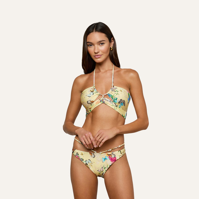 A woman with long brown hair stands against a plain light background, looking at the camera with a relaxed expression while wearing the Sirena Bikini Top—a yellow floral halter style featuring pearl accents.