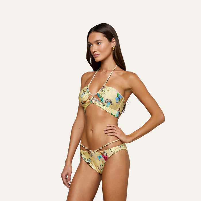 A woman poses against a plain, light background wearing the Sirena Bikini Top, a yellow floral halter bikini with a butterfly print, paired with matching bottoms.