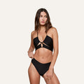 A woman wears the Sirena Halter Bikini Top, featuring a black bohemian style with ruched cups and gold hardware, standing against a plain light background and looking to the side.