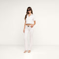 A woman wearing sunglasses, a short-sleeved cropped white top, the Sneeki Low-Rise Straight-Leg Jean in super stretch denim with a raw hem, and heeled sandals stands against a plain white background.