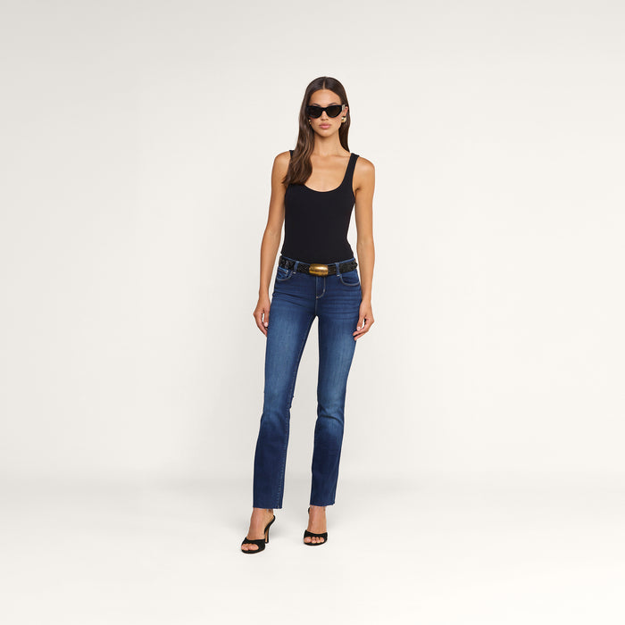 A woman with straight brown hair and a neutral expression, wearing a black tank top, the Sneeki Low-Rise Straight-Leg Jean in dark wash super stretch denim, black sandals, and sunglasses, stands against a plain white background with one hand at her side.