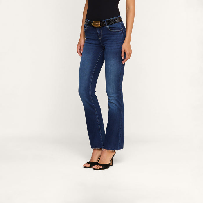 A woman stands against a plain white background wearing the Sneeki Low-Rise Straight-Leg Jean in dark blue super stretch denim with raw hems and subtle fading on the thighs, paired with a black top, black belt, and black open-toe heels.