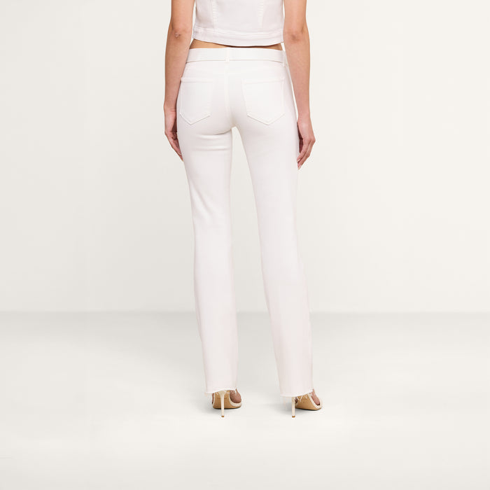 A woman stands facing away, wearing the Sneeki Low-Rise Straight-Leg Jean in white with a raw hem, paired with a white cropped top and beige heels against a plain white background.