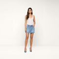A woman with long brown hair and a neutral expression stands against a white background wearing a white tank top, blue heels, and the Solene Jean Mini Skirt—a light wash, all-cotton denim mini skirt with buttons.