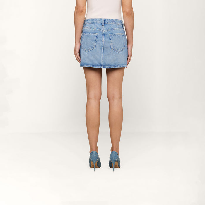 A person stands facing away, wearing the Solene Jean Mini Skirt in a light wash all-cotton denim, paired with matching denim high heels against a plain white background.
