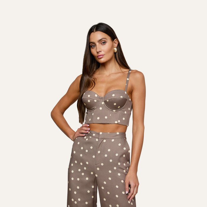 Wearing the Sonya Bustier in taupe with a smocked back, paired with high-waisted cardamom polka dot pants, the woman stands against a plain light background. She has long, straight hair and a neutral expression.