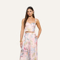 Wearing the Sonya Bustier—a light pink bustier top with spaghetti straps and a delicate palm leaf print—paired with matching wide-leg pants, a woman with long brown hair accessorizes with gold earrings and a gold bracelet as she stands against an off-white background.