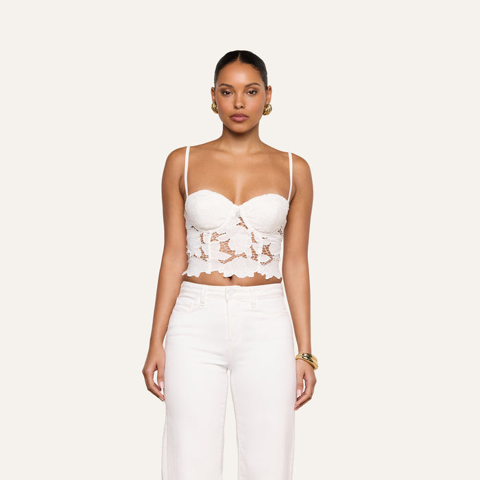 A woman stands against a light background, wearing the Sonya Bustier—a white eyelet crop top with spaghetti straps—paired with white pants. She accessorizes with gold hoop earrings and a gold bracelet, looking directly at the camera.