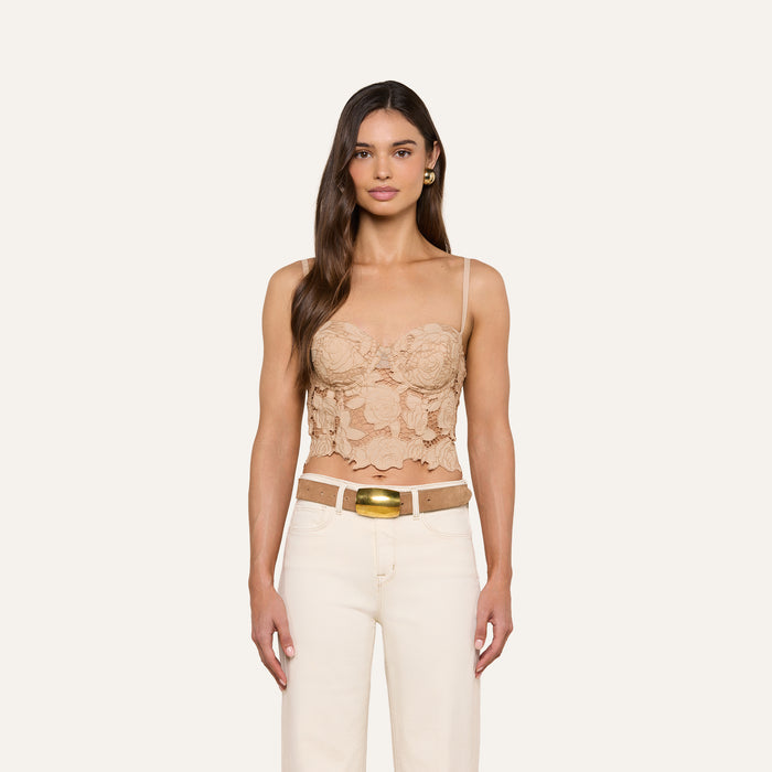 Wearing the Sonya Bustier, a woman with long brown hair stands against a plain light background; she pairs the beige cotton eyelet top with adjustable straps with high-waisted cream pants and a wide tan belt.