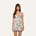 A woman with long brown hair and minimal makeup stands facing forward against a plain, light-colored background. She is wearing the Sonya Bustier—a floral print butterfly bustier top—paired with high-waisted shorts and accessorized with a gold bracelet on her right wrist.