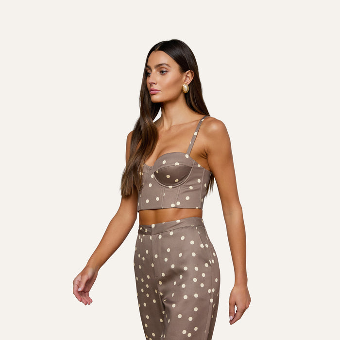 Woman with long brown hair wears the Sonya Bustier, a taupe crop top crafted from lightweight twill and featuring white cardamom polka dots, paired with matching high-waisted pants, standing against a plain light background.