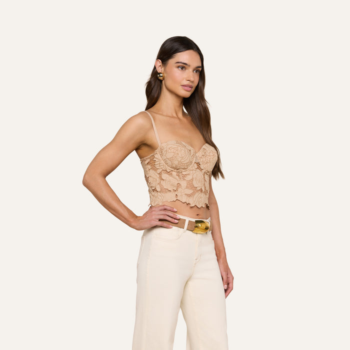 A woman stands in profile against a plain white background, wearing the Sonya Bustier—a beige floral lace top with adjustable straps—paired with cream high-waisted pants and gold hoop earrings. Her long brown hair is loose, and she has a neutral expression.