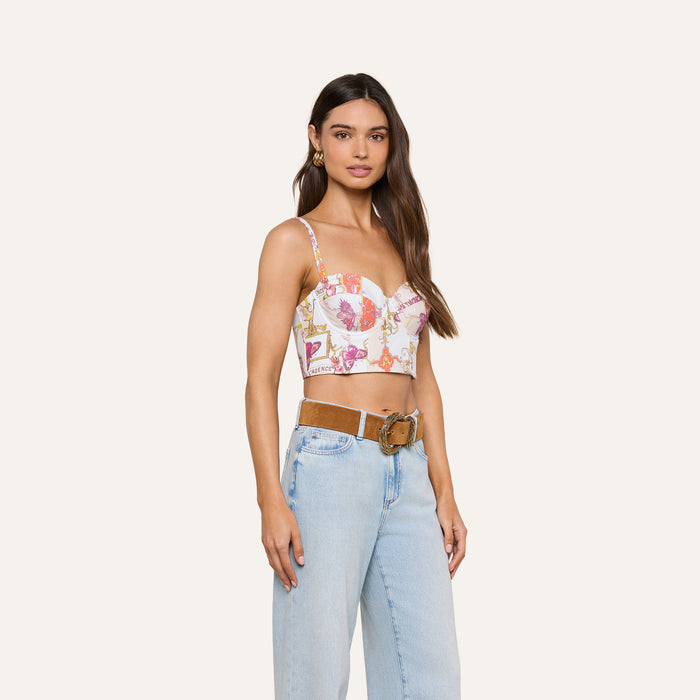 A woman stands and poses against a plain off-white background, wearing the Sonya Bustier—a baroque toile print bustier top with spaghetti straps—paired with light blue high-waisted jeans and a brown belt with a large buckle.