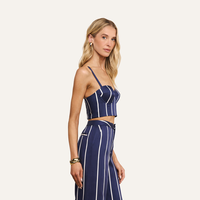 Wearing the Sonya Bustier in a navy stripe, this woman with long blonde hair pairs it with matching high-waisted pants. She stands in profile against a plain light background, accessorized with earrings and a bracelet.